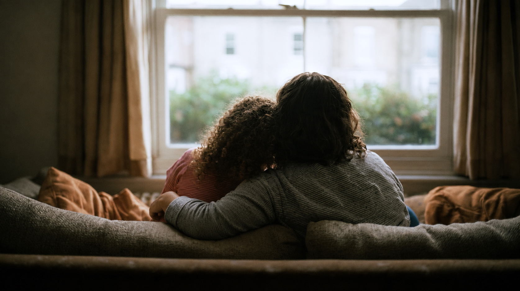 A mother and her daughter sitting close together on a sofa, seen from behind, looking out of a bright window.